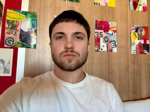 Christian Lotz has a dark brown beard and hair and is wearing a white T-shirt. He is looking directly at the camera. There are six brightly coloured red, and green portrait shaped collage-style artworks on the wall behind him, which has a brown timber-effect.