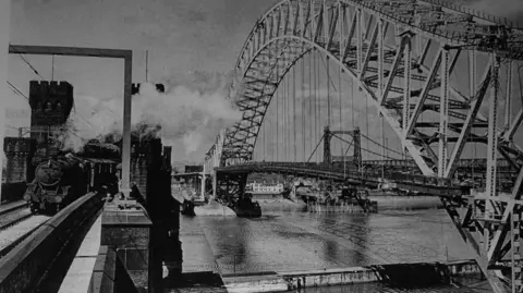 The distinctive metal structure of Runcorn bridge in the early 1960s alongside a steam train