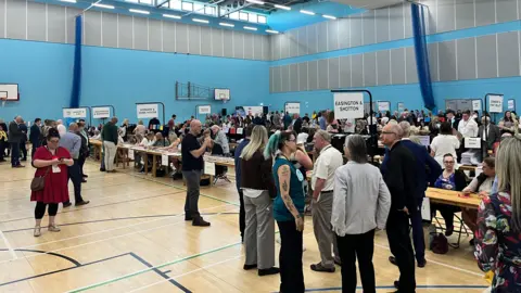 A picture of the count for Durham County Council.  Rows of tables have been laid out in a sports hall where people are seated and counting the votes. The room is filled with people awaiting the result.
