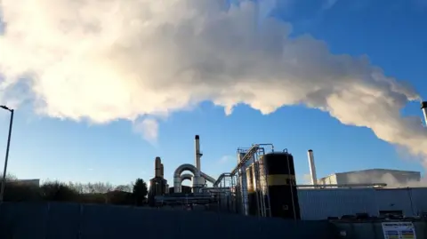 Ummer Daraz A wide shot of a factory plant, with multiple chimneys and metallic industrial structures in view. Plumes of smoke/emissions can be seen coming out of one large chimney and spreading across the skyline.