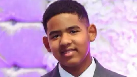 West Midlands Police Alama pictured smiling while wearing a grey suit in front of a purple background. He has short black hair and is clean shaven.