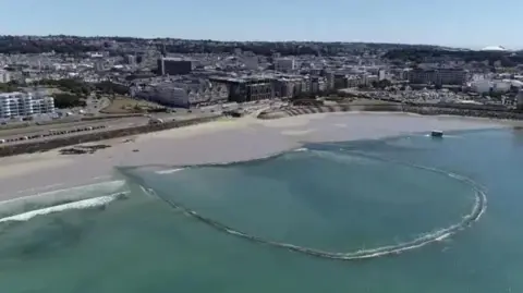 A u-shaped wall encloses sea water off a beach in St Aubin's Bay with a built up St Helier town centre in the background.