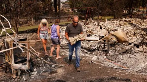 Reuters A man carrying a small statue as he walked through burned-out rubble and ashes, with a child's swing seen in a garden in the background - two women wearing vests and shorts are behind him, one turning to look at the ruins 