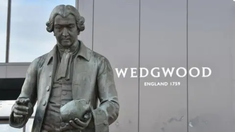 A statue of a man dressed in old-fashioned clothes and holding a pot in his left hand. Behind him is a building which says "Wedgwood - England 1759".