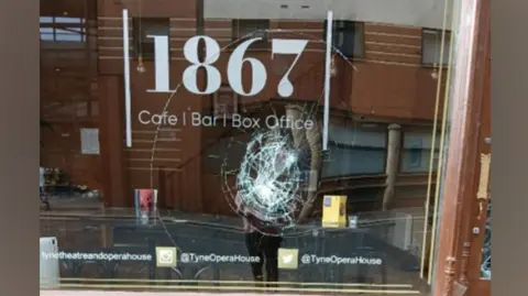 Tyne Theatre & Opera House A glass window with the words '1867 cafe, bar, box office' written on the glass. Underneath is a circular smash in the class with broken glass on the pavement outside.