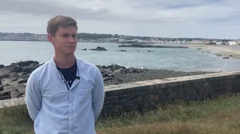 Brandon Ashplant, standing near a rocky shoreline with the sea in the background. He has blonde hair and is wearing a light blue button-up shirt over a dark-coloured shirt.