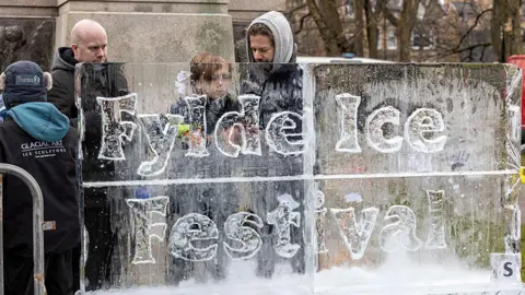 Mark Liebenberg Ice sculptures from Glacial Art stand behind an ice sculpture with the words 'Fylde Ice Festival' engraved in it in Lytham town centre. 