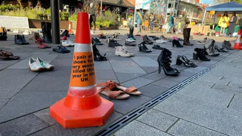 A display of pairs of shoes on a city centre high street. Shoes are lined up in rows and a stall with people around it is in the background. A traffic cone is in the foreground with an orange sign that says "one pair of shoes for every woman killed this year in the UK". 