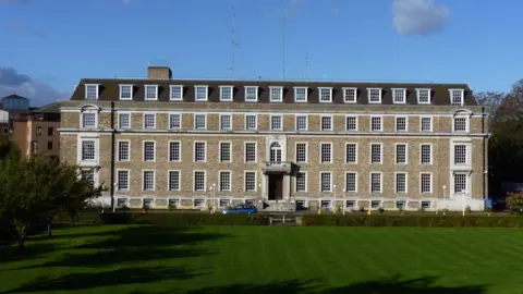 Shire Hall is a large, brick building with four stories and many windows. It is set in a large lawned area with hedges. There is a modern building behind it, to the left of the image.