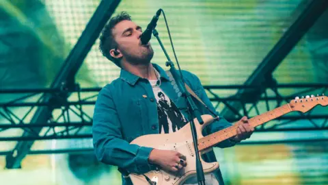 Sam Fender performing on stage at St James' Park. He has short dark hair and a dark stubbly beard, and is wearing a blue denim jacket with a white t shirt sporting black figures on it. He sings into a microphone while holding a guitar. There are green and yellow lights on a screen behind him.
