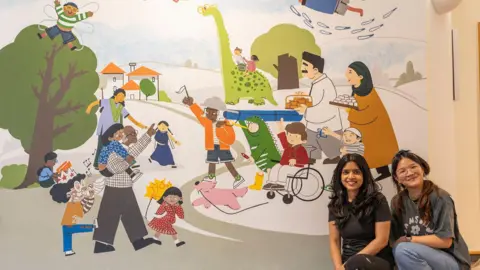 Shooting Star Children's Hospices Two women sitting in front of a colourful mural installed at a children's hospice which features parents, children, a dog and a dinosaur walking around a park. 