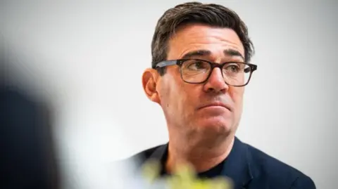 A headshot of Andy Burnham, wearing a black suit over a black t-shirt and black spectacles. He has a serious expression on his face.