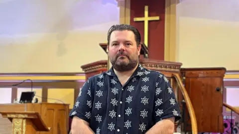 Jonathan stands inside a church in front of a wooden pulpit with a gold cross on a wooden backdrop.