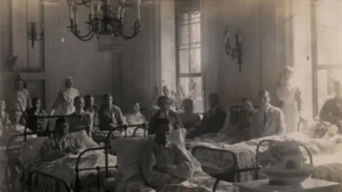 North Lincolnshire Museums A chandelier hangs from the ceiling in a ballroom that has been transformed into a makeshift hospital ward. Soldier sit up in bed, arms crossed, watched by nurses.