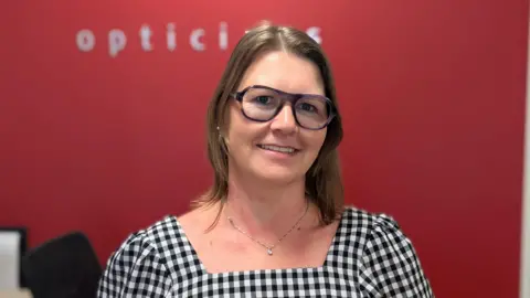 Diane Faulkner is standing in front of a red painted wall, with a singing spelling opticians on it. She is wearing a checked dress, has shoulder length brown hair and is wearing glasses.
