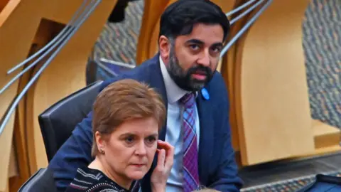 Getty Images Nicola Sturgeon yang duduk di depan Humza Yousaf adalah tempat duduk mereka di Kamar Parlemen Skotlandia. Sturgeon menyentuh pipi kirinya dengan tangan kanannya dan mengenakan lingkaran putih kemeja hitam. Yousaf melihat ke arah kamera dan mengenakan setelan biru, dasi putih, dan dasi tartan ungu dan biru. 