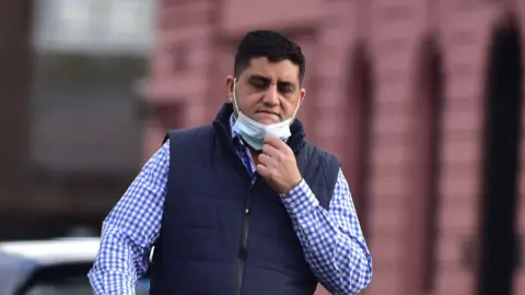 A dark haired man in a check shirt and gilet with a facemask pulled down on his chin