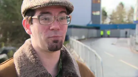 A man wearing glasses and a flat cap, and a brown fur-lined collar jacket, looks to the right of the camera, as he stands outside of a hospital