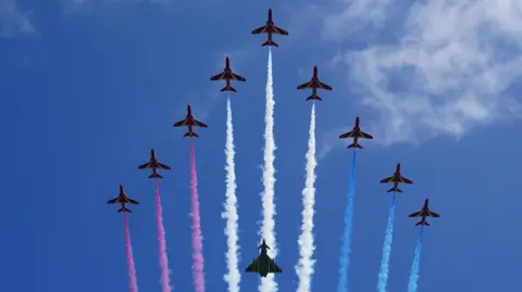 Nine Red Arrow jets and a Typhoon FGR4 aircraft perform a flypast. The Red Arrow jets have red, white and blue smoke coming out of their rears. The Red Arrows and in a V formation with the Typhoon FGR4 positioned underneath them. It is a sunny day with thin cloud cover in the sky.