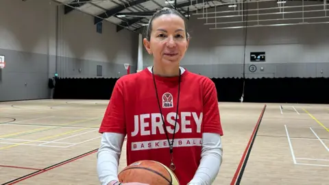 Timea Kraft stands in the middle of an indoor sports hall holding a basketball with both hands. She is wearing a red T‑shirt with the text “JERSEY BASKETBALL” and a JBBA logo above it, along with a white long‑sleeved top underneath. The sports hall features marked courts, high ceilings with lighting rigs, and a scoreboard on the far wall.