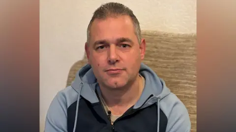 A man in a light blue tracksuit top with short hair sitting on a beige sofa looks at the camera unsmilingly 