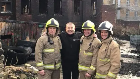 Supplied A group of firefighters stand in front of a burned building. They wear beige protective gear with fluorescent stripes, and yellow helmets.