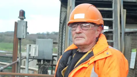 Jamie Niblock/BBC Malcolm Stevens standing outside on a high metal walkway at a sugar beet factory. He is wearing an orange hard hat and an orange coat. He has clear safety goggles on over his glasses and is looking into the distance. 