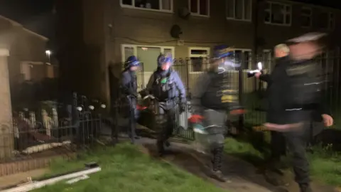 Photograph of the police taking part in a raid on the Hart Mill Estate in Mossley. The image is blurred to protect the identity of the officers. 