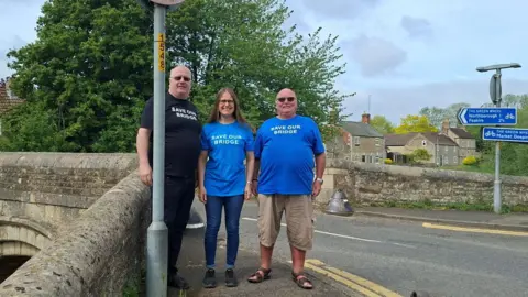 Paul Frith Two men and a woman standing at the entrance to a narrow stone bridge while wearing blue T-shirts with a slogan reading "Save our bridge". Trees and houses can be seen in the background.