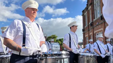 A group of men in white shirts and white gloves carry drums 