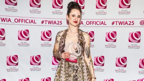 The picture shows Holly Miller holding her award at the Wedding Industry Awards. Holly is wearing a bright patterned dress and smiling at the camera.