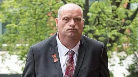 A man wearing a white shirt, a grey suit jacket and a red patterned tie with a red ribbon on his jacket faces the camera. There are trees in the background.