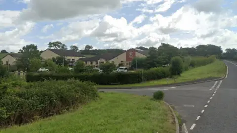 Google Maps A large beige single-storey brick building, with a brown roof, sits behind a low green hedge. In front of the hedge is a thin strip of grass and a black tarmac road. To the right of the building is another larger road, which runs up the side of it. 