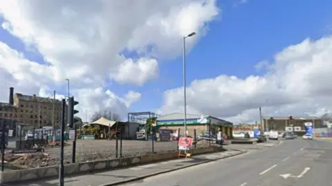 A fenced off plot of land on an urban road. Street furniture include traffic lights and lamp-posts can be seen.