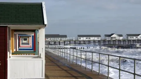 A series of colourful beach huts on the promenade in Southwold, with the huts on the left of the picture, the sea and waves crashing on the right and a pier in the distance.