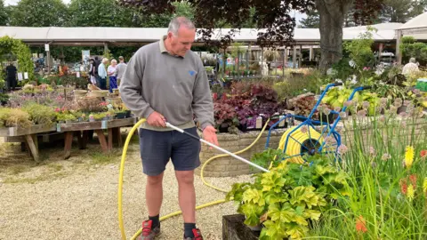 A man in a grey jumper and blue shorts waters plants using a long yellow hose.