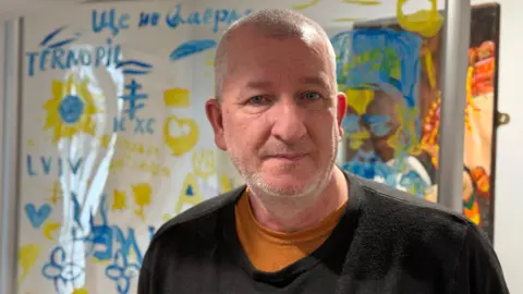 A balding man with grey stubble wearing a black sweatshirt and looking straight at the camera. There's a blue and yellow mural on the wall behind him.