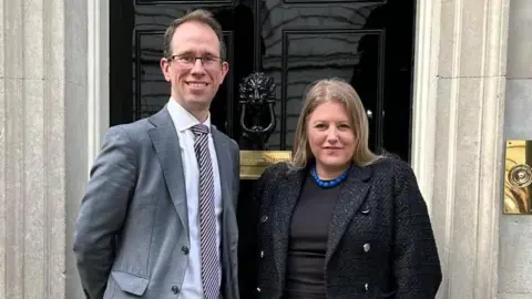 Matthew Barber and Donna Jones stand outside 10 Downing Street. Mr Barber is taller, has receding brown hair, and wears a light grey suit and glasses. Ms Jones has long, straight, fair hair, and wears a black jacket over a black top with a blue beaded necklace.