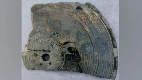 About a quarter of a round 17th Century wooden lantern base. In the middle is a large hole, surrounded by five smaller holes. The hole is surrounded by concentric circles until it reaches its raised edge. 