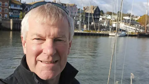 Man smiles in front of a pier with boats and houses in the distance.