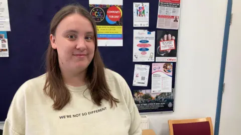 James Grant/BBC Holly Sampson with long brown hair and a "we're not so different" top in front of a noticeboard