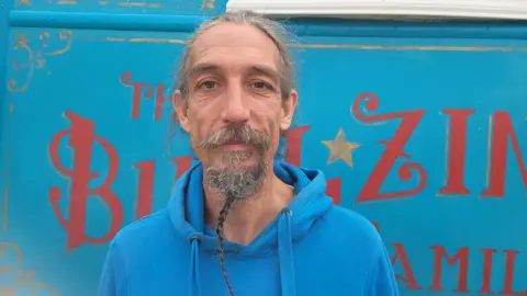 Chris Bullzini is standing in front of a blue circus wagon. He is wearing a blue hoodie and is smiling.