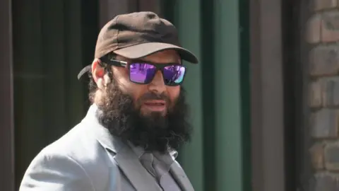 Close-up image of Farris Wilson with a full dark beard wearing a light grey blazer over a light shirt, a dark baseball cap and mirrored purple sunglasses. He is standing outside in bright sunlight beside a brick wall and dark green window frames.