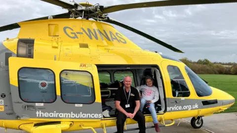 Air Ambulance Service A man in a black t-shirt and a girl in a grey jumper sit in a yellow helicopter which is grounded.