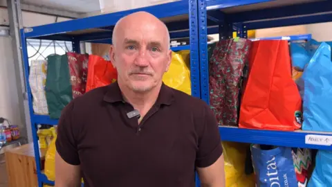 Former champion boxer Barry McGuigan wears a burgundy polo shirt and faces camera with bags of food at the bank behind him