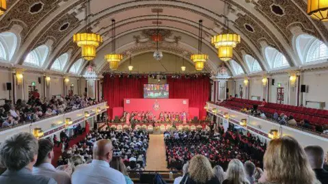 Sophie Dallaway A large room with an ornate ceiling with lights hanging from the ceiling with a red backdrop at the end of the room with red seats lining the balcony with stalls and people sitting watching a graduation ceremony.