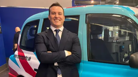 BBC James McMurdock is standing in front of a Hackney cab carriage, which is painted in the colours of Reform UK (blue, white and red). He is wearing a navy blue suite and a blue tie.