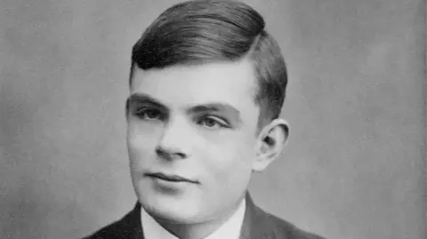 Getty Images A black and white photograph of Alan Turing who has short hair. He is sitting in front of a backdrop and is wearing a white shirt and blazer.