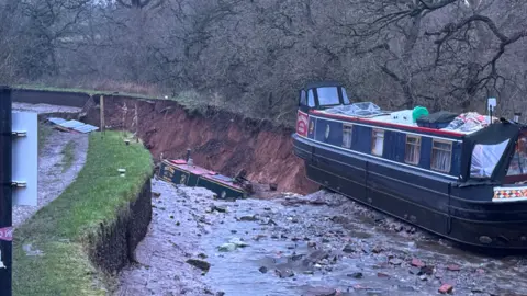 Shropshire Fire and Rescue Service Two barges at the scene of a canal sinkhole. Trees are on one side of the canal.