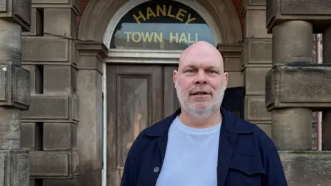 Simon Edwards, a bald man with a grey beard, wearing a navy blue jacket over a white t-shirt. He is standing outside a stone building which has the words Hanley Town Hall in gold lettering on a arched window above a wooden door.